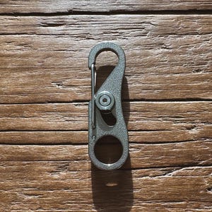 May include: A silver-colored carabiner clip with a spring-loaded gate and a circular opening at each end. The clip has a textured surface and a central pivot point. The background is a wooden surface.