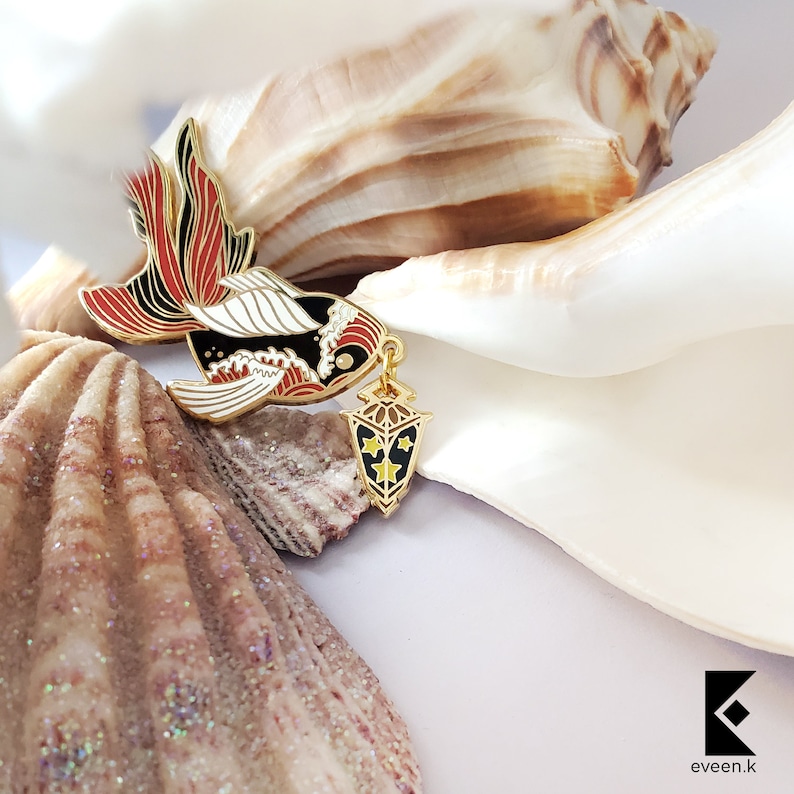 May include: A gold-colored enamel pin in the shape of a fish with red, black, and white details. The pin has a dangling lantern charm with yellow stars and a black background. The pin is set against a backdrop of seashells.