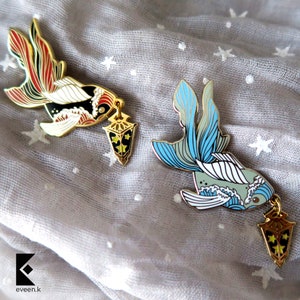 May include: Two enamel pins featuring goldfish designs with intricate details. One pin has red, black, and gold accents, while the other showcases blue and gray tones. Each fish holds a gold lantern charm with star details.