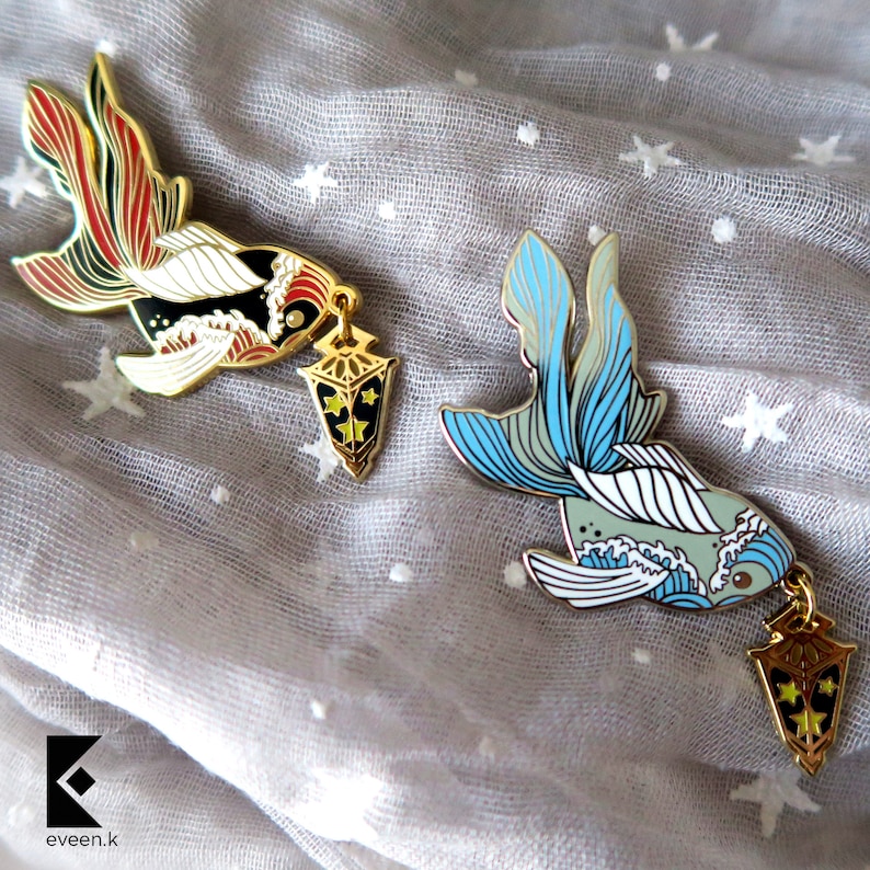 May include: Two enamel pins featuring goldfish designs with intricate details. One pin has red, black, and gold accents, while the other showcases blue and gray tones. Each fish holds a gold lantern charm with star details.