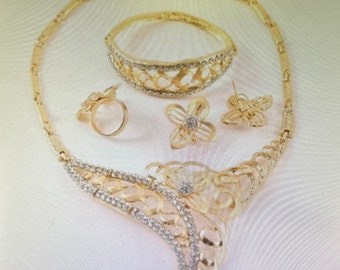 May include: A gold-toned jewelry set featuring a necklace, bracelet, ring, and earrings. The necklace and bracelet have a decorative openwork design with clear rhinestones. The ring and earrings are flower-shaped with a central rhinestone.