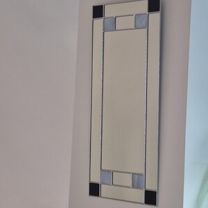 May include: A rectangular wall mirror with a cream-coloured reflective surface. The mirror has a silver border and decorative black and light blue square accents at the top and bottom. It is mounted on a light beige wall.