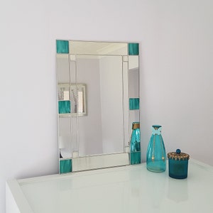 May include: A rectangular mirror with a silver frame and teal glass accents. The mirror is sitting on a white countertop with a teal glass bottle and a teal glass jar.