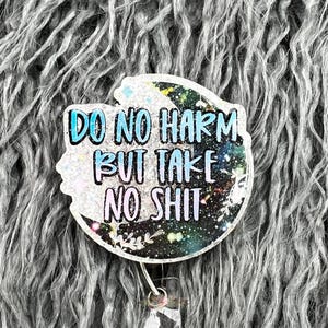 以下が含まれることがあります： 「DO NO HARM BUT TAKE NO SHIT」の文字が青と白で書かれた、白くキラキラ光るバッジリール。 バッジリールは銀河をテーマにしたデザインで、透明なプラスチッククリップが付いています。 背景はグレーのふわふわした素材です。