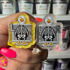 Op de afbeelding: Twee acryl badges met een cartoon eend met een skeletafbeelding binnen een röntgenfoto-kader. De ene badge heeft een gele glitterafwerking, de andere een heldere iriserende glitterafwerking. Beide tonen de letters L en R.