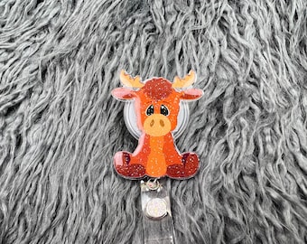 Può includere: Un portabadge a forma di alce arancione e rosso, stravagante, con accenti glitterati. L'alce ha grandi occhi espressivi ed è seduto. Il portabadge è attaccato a un cordino retrattile di plastica trasparente e a un bottone decorativo glitterato.