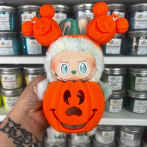 May include: A whimsical Halloween-themed figurine featuring a smiling orange pumpkin head and a character wearing a white and green furry hood. The figurine has orange pumpkin ears and the text "THE MONS" on a green band.
