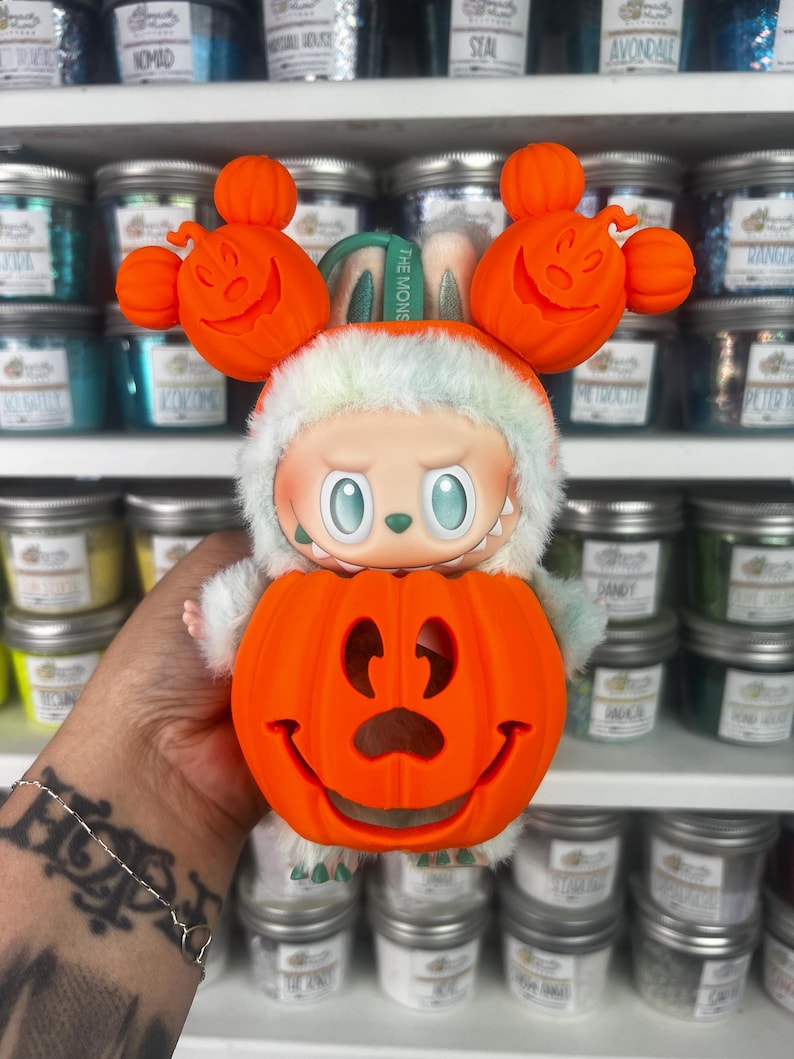 May include: A whimsical Halloween-themed figurine featuring a smiling orange pumpkin head and a character wearing a white and green furry hood. The figurine has orange pumpkin ears and the text "THE MONS" on a green band.
