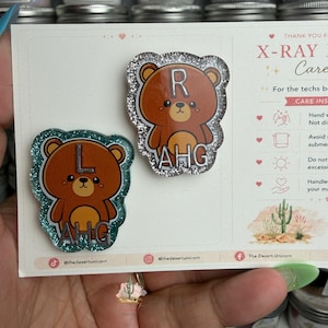 Può includere: Due badge a forma di orso glitterati. Uno è bordato di glitter argentati e ha la lettera "R" e le lettere "AHG". L'altro è bordato di glitter verde acqua e ha la lettera "L" e le lettere "AHG". I badge sono su una carta bianca.