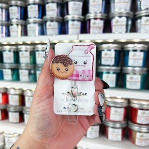 Puede incluir: Un cartón de leche rosa y blanco con una cara sonriente y una galleta marrón y blanca con una cara sonriente. El cartón de leche dice "MILK". El charm está unido a un portagajes retráctil plateado. El portagajes tiene una cubierta de plástico transparente y un clip plateado. El charm está sobre un fondo blanco con el texto "The Dysfunctional Unicorn" y una dirección de sitio web.