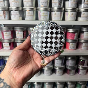 May include: A circular badge reel featuring a black and white checkered pattern in the center, surrounded by a border of silver and black glitter. A small, three-dimensional cactus charm hangs from the bottom of the circular badge reel.