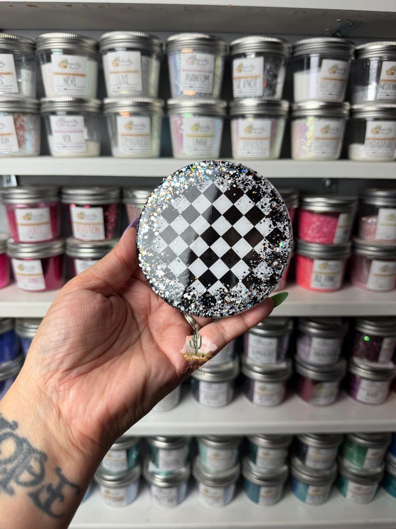 May include: A circular badge reel featuring a black and white checkered pattern in the center, surrounded by a border of silver and black glitter. A small, three-dimensional cactus charm hangs from the bottom of the circular badge reel.