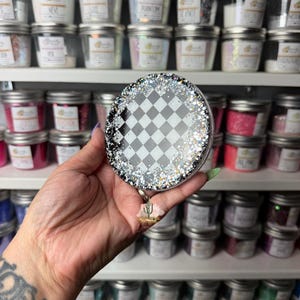 May include: A circular badge reel featuring a black and white checkered pattern in the center, surrounded by a border of chunky silver and black glitter. A small, three-dimensional cactus charm hangs from the bottom of the circular housing.
