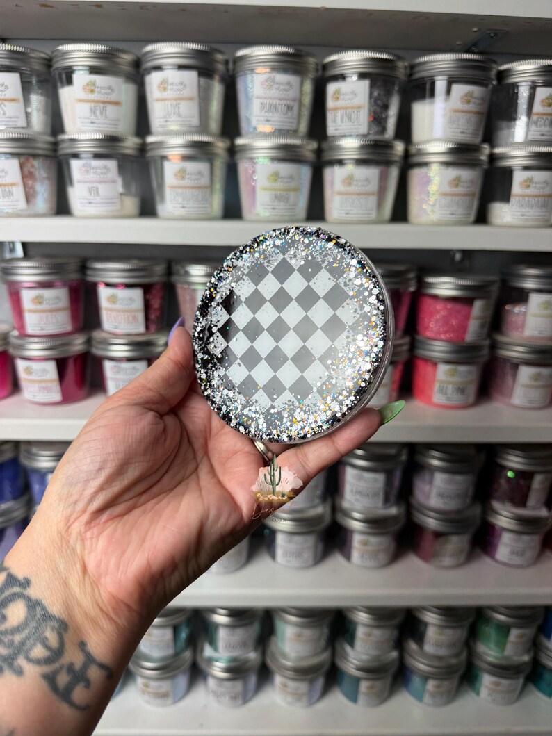 May include: A circular badge reel featuring a black and white checkered pattern in the center, surrounded by a border of chunky silver and black glitter. A small, three-dimensional cactus charm hangs from the bottom of the circular housing.