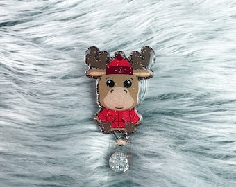 Può includere: Un simpatico porta badge a forma di alce con cappello e giacca rossi. L'alce ha corna marroni e una finitura glitterata. Il porta badge ha una base in plastica trasparente e un bottone decorativo e scintillante.