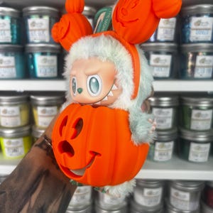 May include: A whimsical Halloween-themed figurine featuring a character with a light green body and a pumpkin head. The figurine is wearing an orange headband with Mickey Mouse-shaped pumpkins. The character is inside a carved orange pumpkin.