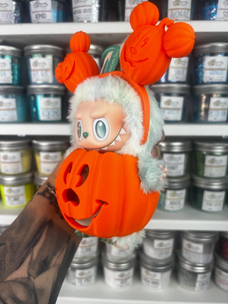 May include: A whimsical Halloween-themed figurine featuring a character with a light green body and a pumpkin head. The figurine is wearing an orange headband with Mickey Mouse-shaped pumpkins. The character is inside a carved orange pumpkin.