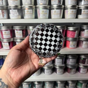 May include: A circular badge reel featuring a black and white checkered pattern in the center, surrounded by a border of clear resin embedded with silver and black glitter. A small cactus charm hangs from the bottom of the circular badge reel.