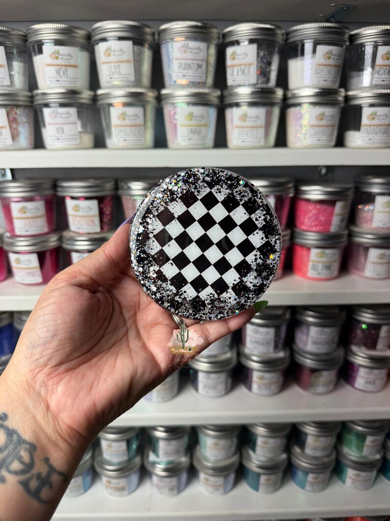 May include: A circular badge reel featuring a black and white checkered pattern in the center, surrounded by a border of clear resin embedded with silver and black glitter. A small cactus charm hangs from the bottom of the circular badge reel.