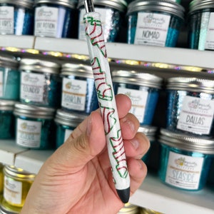 May include: A white pen with red and green accents and the word "Dallas" printed on it. The pen has a silver tip and a black writing end. The background features jars of glitter.