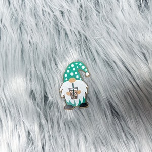 May include: A green and white gnome with a white polka dot hat holding a green and brown boba tea cup. The gnome is standing on a white fluffy surface.