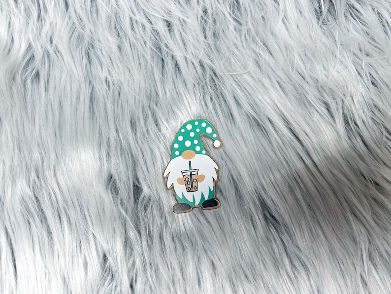 May include: A green and white gnome with a white polka dot hat holding a green and brown boba tea cup. The gnome is standing on a white fluffy surface.