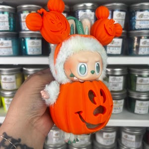 May include: A Halloween-themed figurine featuring a character in a pumpkin costume with Mickey Mouse-inspired ears. The figurine has a white furry trim and a smiling jack-o'-lantern face. The figurine is orange, white, and green.