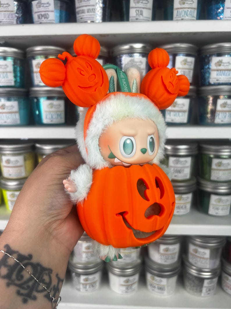 May include: A Halloween-themed figurine featuring a character in a pumpkin costume with Mickey Mouse-inspired ears. The figurine has a white furry trim and a smiling jack-o'-lantern face. The figurine is orange, white, and green.