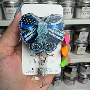May include: A badge reel featuring a blue and black bow design, a police badge, and handcuffs. The reel has a retractable cord and a decorative gem. Jars of glitter are in the background.