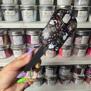 May include: A black spatula featuring a repeating pattern of white and grey robot characters, small orange stars, and pink hearts. The handle is solid black, and the spatula head has a distinct U-shaped cutout at the top edge for hanging storage.