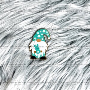 May include: A turquoise gnome with a white hat and white flowers. The gnome is holding a green bunny in its hands. The gnome is standing on a white fluffy surface.