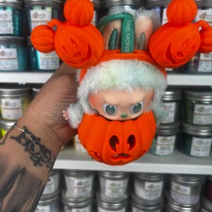 May include: A Halloween-themed collectible figure featuring a character inside an orange pumpkin. The figure has a white and green furry hat and is surrounded by orange pumpkin decorations. The tag reads "THE MONS".