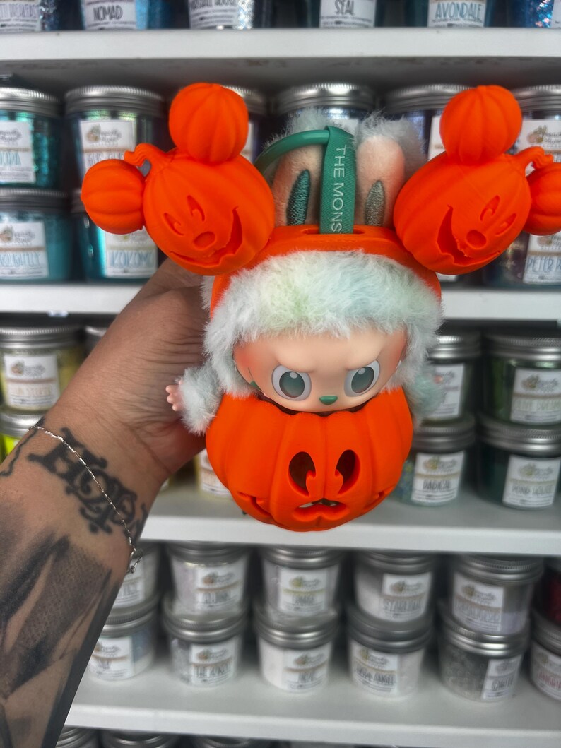 May include: A Halloween-themed collectible figure featuring a character inside an orange pumpkin. The figure has a white and green furry hat and is surrounded by orange pumpkin decorations. The tag reads "THE MONS".