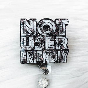 May include: A black and white badge holder with the text "NOT USER FRIENDLY" in white letters with a floral pattern. The badge holder is attached to a clear retractable badge reel with a silver clip.