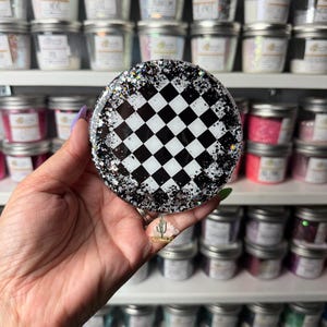 May include: A circular badge reel featuring a black and white checkered pattern in the center, surrounded by a border of black and silver glitter. The design is encased in a smooth, glossy resin finish, creating a reflective and textured appearance.