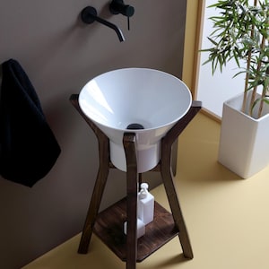 Peut inclure: Un lavabo en céramique blanche avec une large ouverture évasée est posé sur un piétement en bois brun foncé avec une étagère. Le lavabo est monté sur un piédestal à quatre pieds. Le piétement a une étagère de rangement.