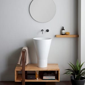 Peut inclure: Un lavabo en céramique blanche avec un design arrondi et évasé est posé sur une étagère en bois avec un porte-serviettes. L'étagère comporte deux niveaux et est soutenue par un seul poteau en bois épais. Le lavabo se trouve dans une salle de bain moderne avec un miroir rond et une étagère en bois au-dessus du lavabo.