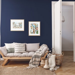 Puede incluir: Una sala de estar con un sof&aacute; fut&oacute;n gris claro con dos almohadas y una manta de lana blanca y negra. El fut&oacute;n est&aacute; frente a una pared azul oscuro con dos cuadros enmarcados. Un taburete blanco con una cesta de lana est&aacute; en la esquina de la habitaci&oacute;n. Una puerta blanca est&aacute; abierta a otra habitaci&oacute;n. El suelo es de madera marr&oacute;n claro.