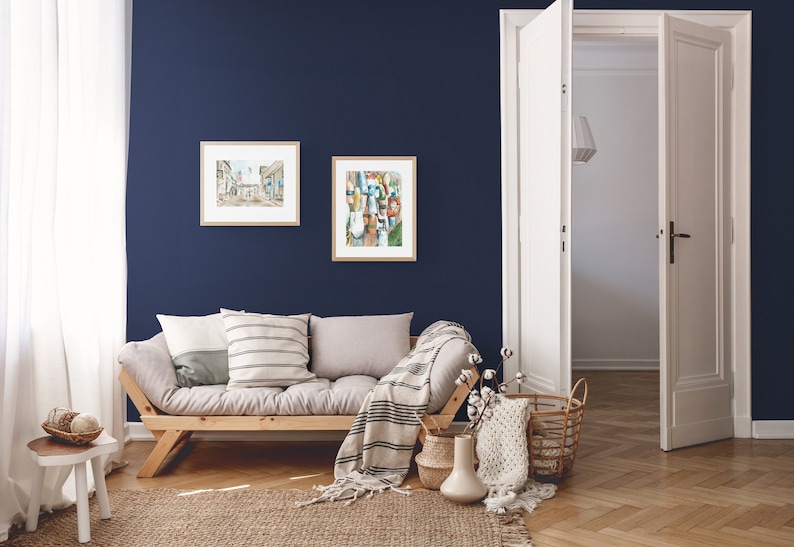 Puede incluir: Una sala de estar con un sof&aacute; fut&oacute;n gris claro con dos almohadas y una manta de lana blanca y negra. El fut&oacute;n est&aacute; frente a una pared azul oscuro con dos cuadros enmarcados. Un taburete blanco con una cesta de lana est&aacute; en la esquina de la habitaci&oacute;n. Una puerta blanca est&aacute; abierta a otra habitaci&oacute;n. El suelo es de madera marr&oacute;n claro.