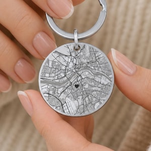 May include: A circular silver-toned metal keychain featuring a detailed black and white street map engraving. The center of the map displays a small, solid black heart icon. The keychain is attached to a standard round metal split ring.