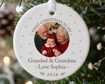 May include: A round white ceramic Christmas ornament featuring a central circular photograph of a baby with grandparents. The text reads FIRST CHRISTMAS AS MY Grandad & Grandma Love Sophia 2026. It is suspended by a white ribbon.