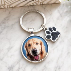 May include: A silver-toned metal keyring featuring a circular pendant with a photographic portrait of a golden retriever dog against a blue background, accompanied by a small, black, textured paw print charm attached to the split ring.