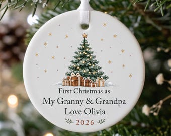 May include: A round white ceramic ornament hanging from a white ribbon. The center features a decorated Christmas tree with wrapped gifts, surrounded by gold stars. Text reads: First Christmas as My Granny & Grandpa Love Olivia 2026.