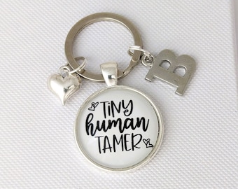 May include: A silver-toned metal keychain featuring a circular pendant with a white background and the black text Tiny human TAMER, accompanied by a small silver heart charm and a silver letter B charm attached to a circular split ring.