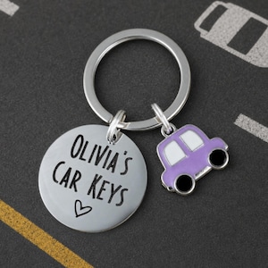 Könnte beinhalten: Ein Schlüsselanhänger aus silberfarbenem Metall mit einer runden Scheibe, die den schwarzen Schriftzug 'OLIVIA'S CAR KEYS' und ein kleines Herz zeigt, ergänzt durch einen Anhänger in Form eines lila-weißen Autos mit schwarzen Rädern.
