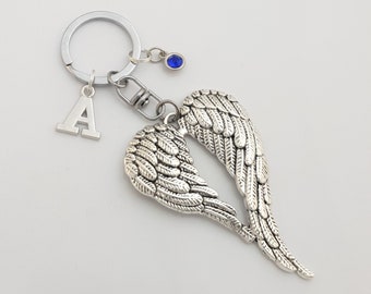 May include: A silver-toned metal keychain featuring a split ring, a lobster clasp, a letter A charm, a small round blue rhinestone charm, and a pair of detailed angel wings. The wings are textured with individual feather patterns in a metallic finish.