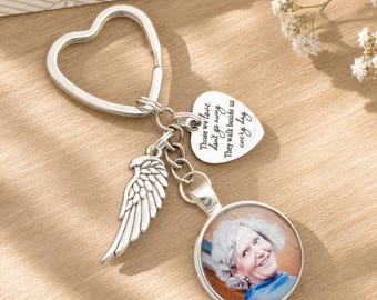 May include: A silver-toned metal keychain featuring a heart-shaped ring, a wing charm, and a heart-shaped pendant engraved with the text "Those we love don't go away, They walk beside us every day." A circular charm displays a portrait of a person.