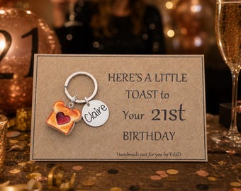 May include: A silver-toned metal keychain featuring a toast-shaped charm with a red heart center and a round disc engraved with the name Claire, attached to a rectangular card with the text: HERE'S A LITTLE TOAST to Your 21st BIRTHDAY.