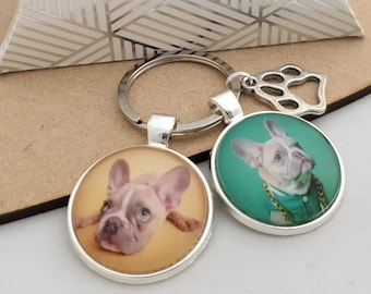 May include: Two silver keychains with a paw print charm. Each keychain has a round glass charm with a different image of a French bulldog. One charm has a green background and the other has a yellow background.