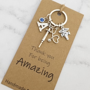 May include: A silver-toned keyring featuring a letter A charm, a blue crystal bead, a syringe charm, a stethoscope charm, and an RN caduceus charm. The keyring is attached to a rectangular brown card printed with the text Thank you For being AMAZING.